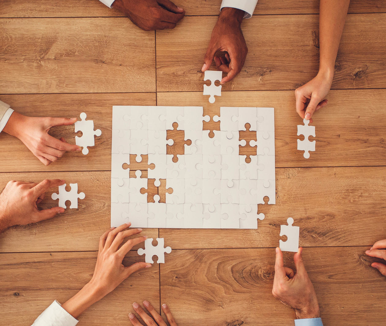 A white jigsaw puzzle with missing pieces being placed into it by a number of different hands. Each hand is holding on to one of the 6 missing pieces