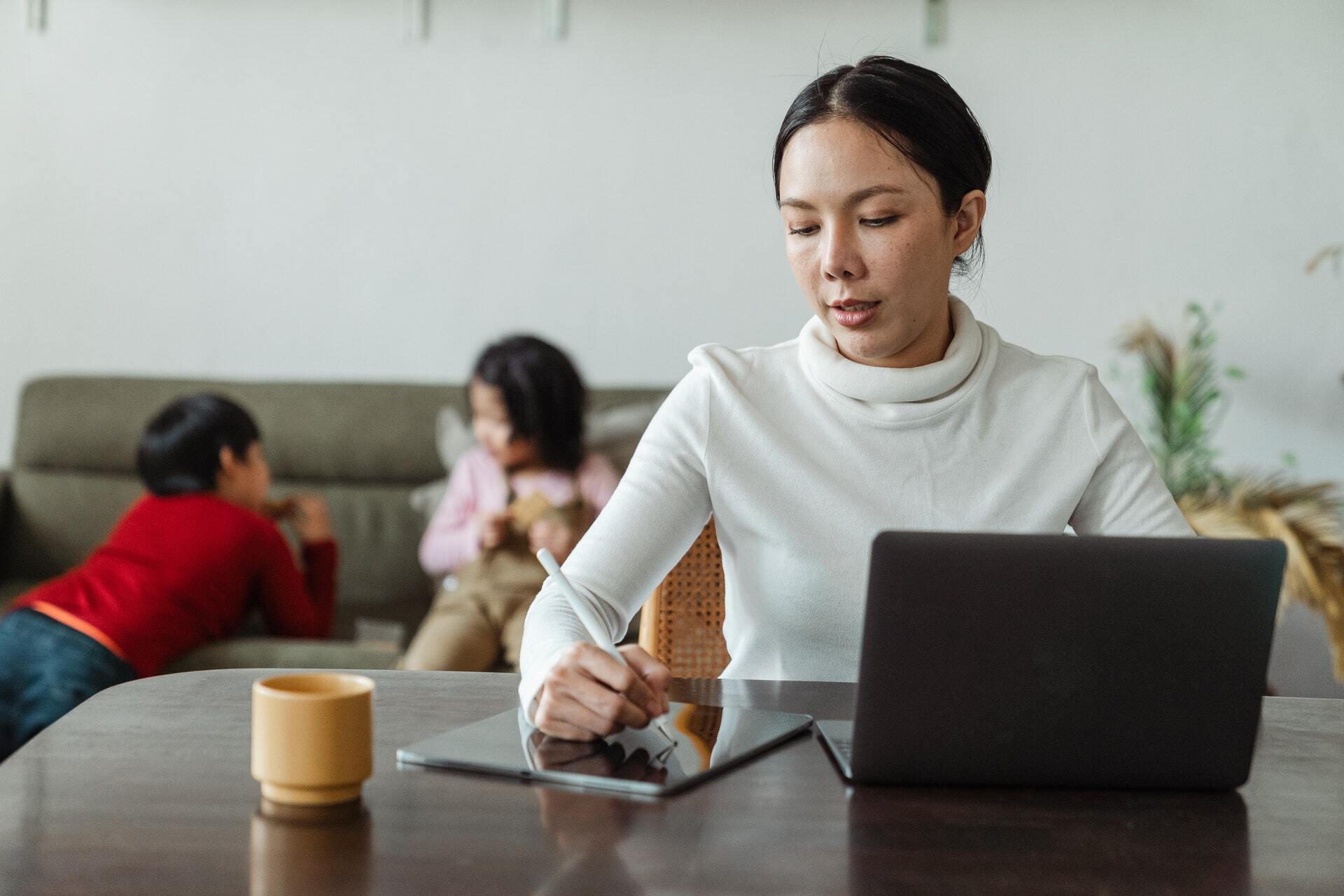 Working mother using tablet and laptop at home with playing kids on background