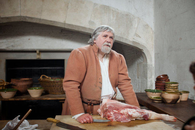 A photograph of a reenactment in a kitchen, with a cook standing next to a leg of pork.