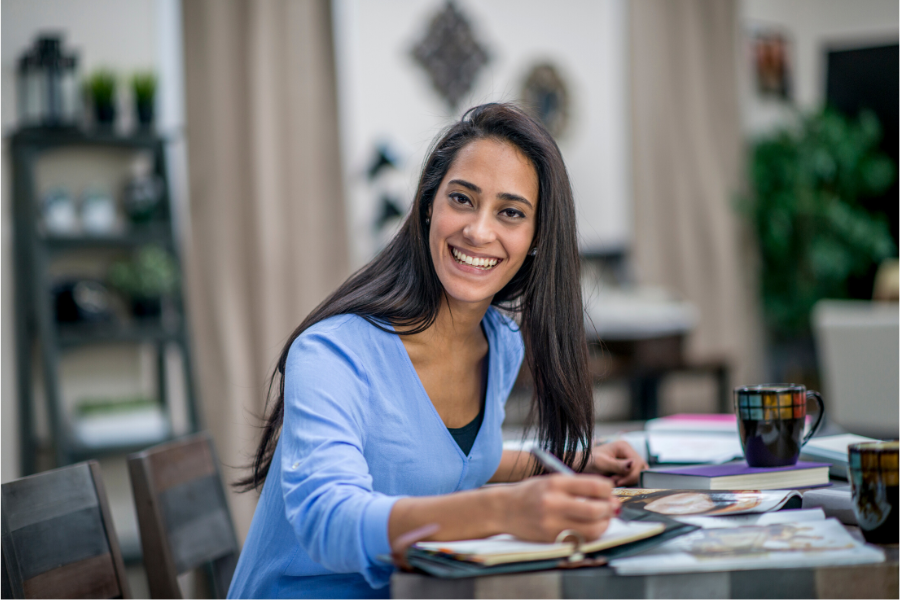 Smiling woman with long dark hair seated at a desk covered with papers, an open diary, books and coffee mugs.