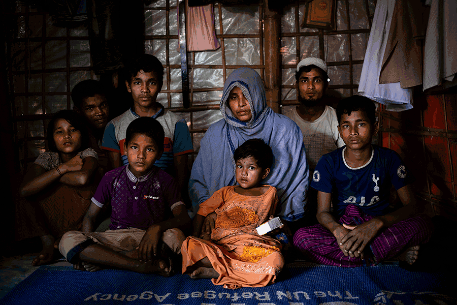 A family of eight sits on a blue UNHCR mat against a wooden wall covered with plastic. Scattered pieces of clothing hang from the walls. The youngest child sitting in the middle holds a box of medicine.