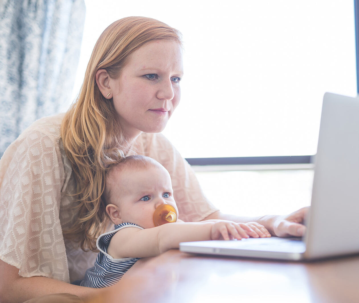 A mother works on her computer while holding her infant baby in her lap.