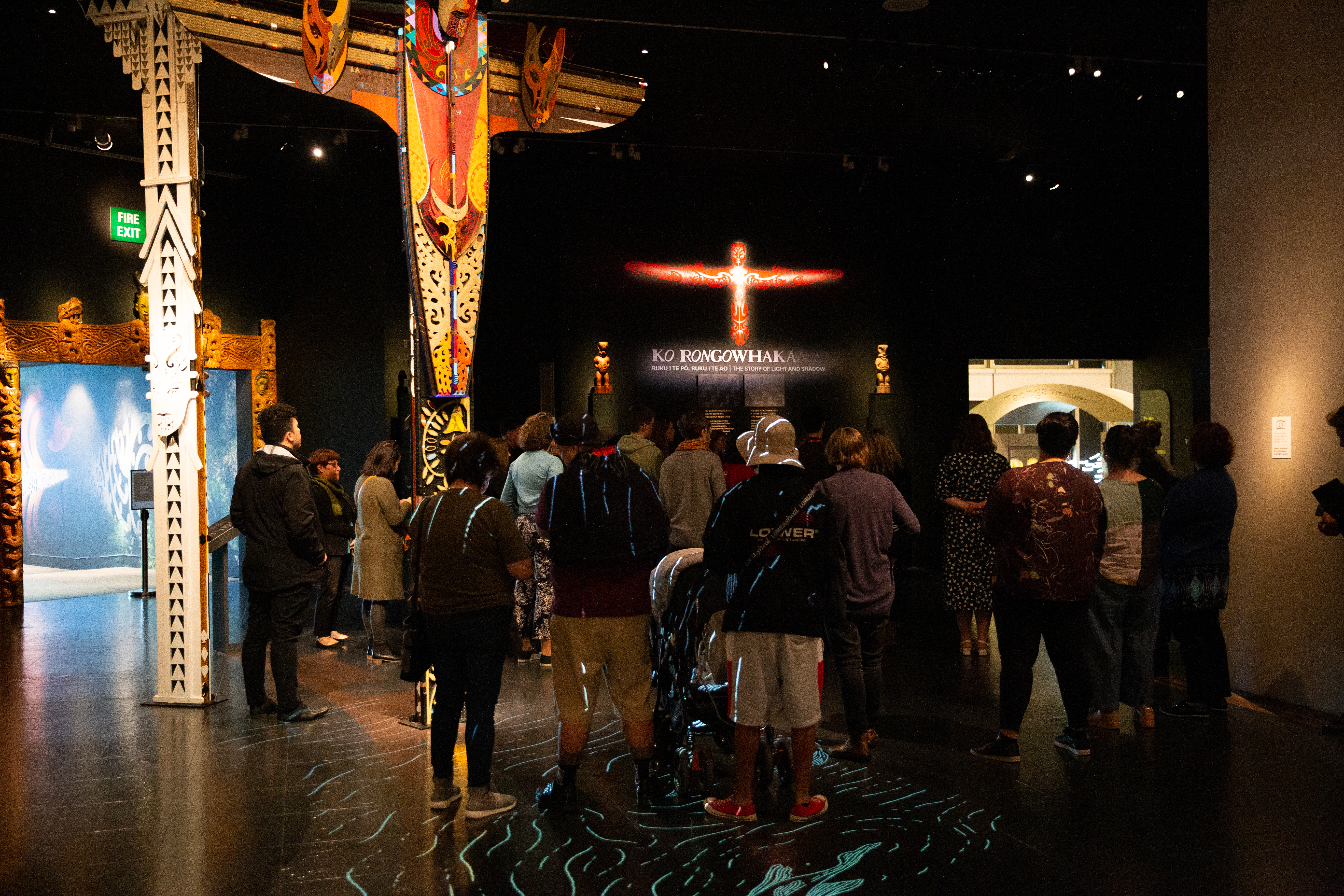 A crowd of people stand in a dark space next to lit up arches rickly carved. They stand before an information panel crowned by a lit up symbol. The symbol is like a red Māori tiki, humanoid but with stylized wings.