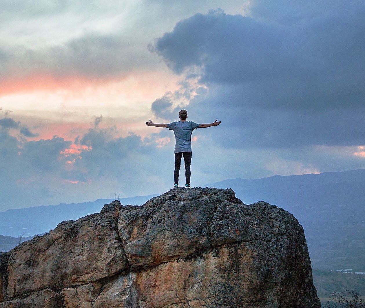 Person standing on a large rock, with arms outstretched, in front of a wide open landscape and cloudy sky.