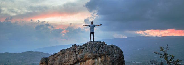 Person standing on a large rock, with arms outstretched, in front of a wide open landscape and cloudy sky.