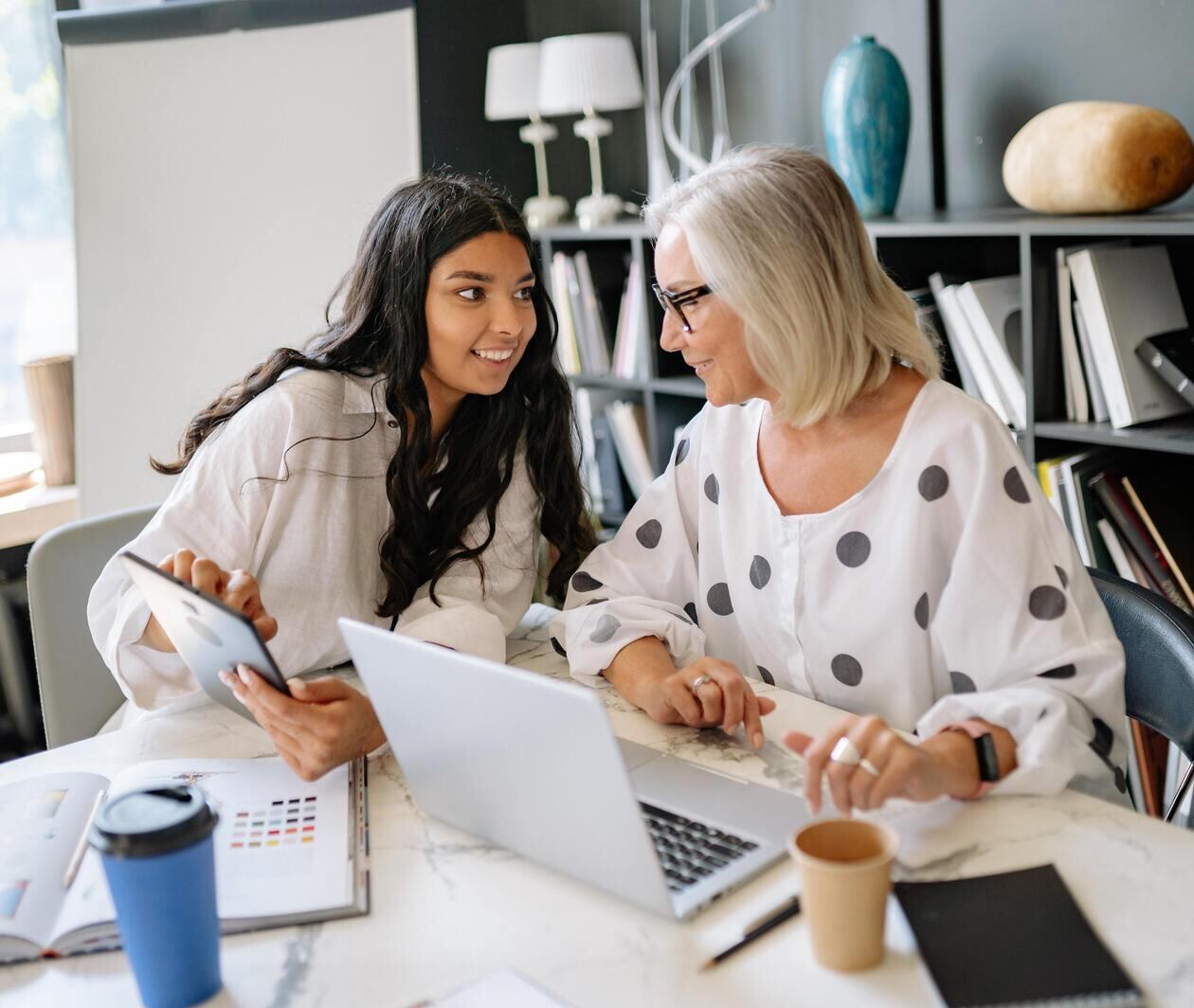 Two women sitting at a desk discussing work on a laptop and tablet
