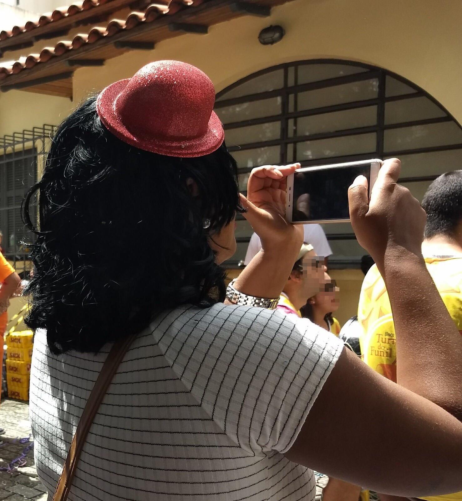 People taking photos using their smartphones during the Carnival in Brazil, 2018. We can see crowds gathered around looking at something, many wearing yellow. In the foreground we can see a woman taking a photo with her smartphone. She wears a red hat.