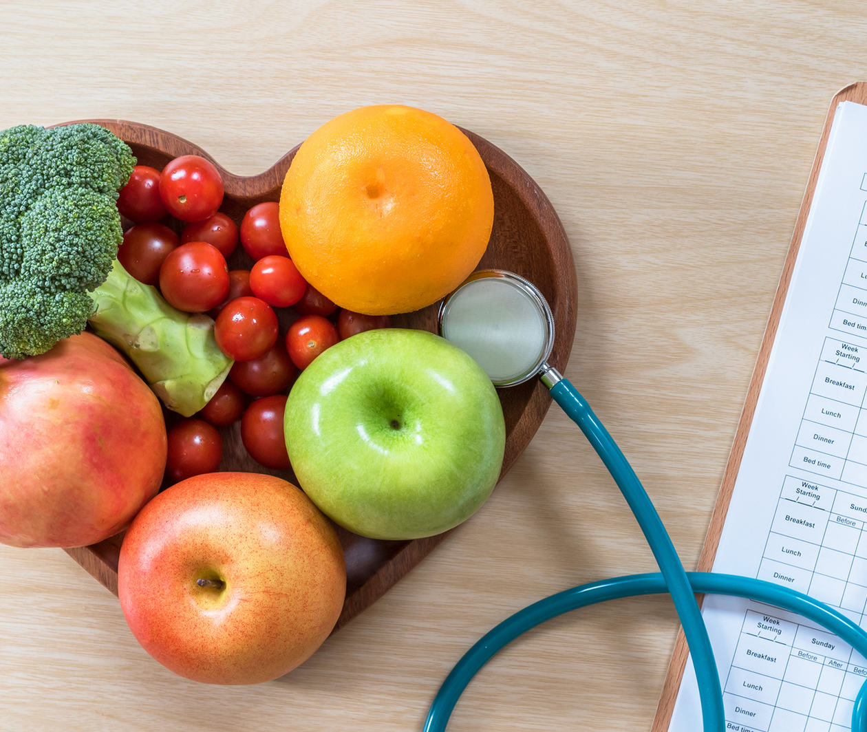 Heart-shaped wooden bowl full of fruit. Sitting on a wooden table next to a stethoscope. 