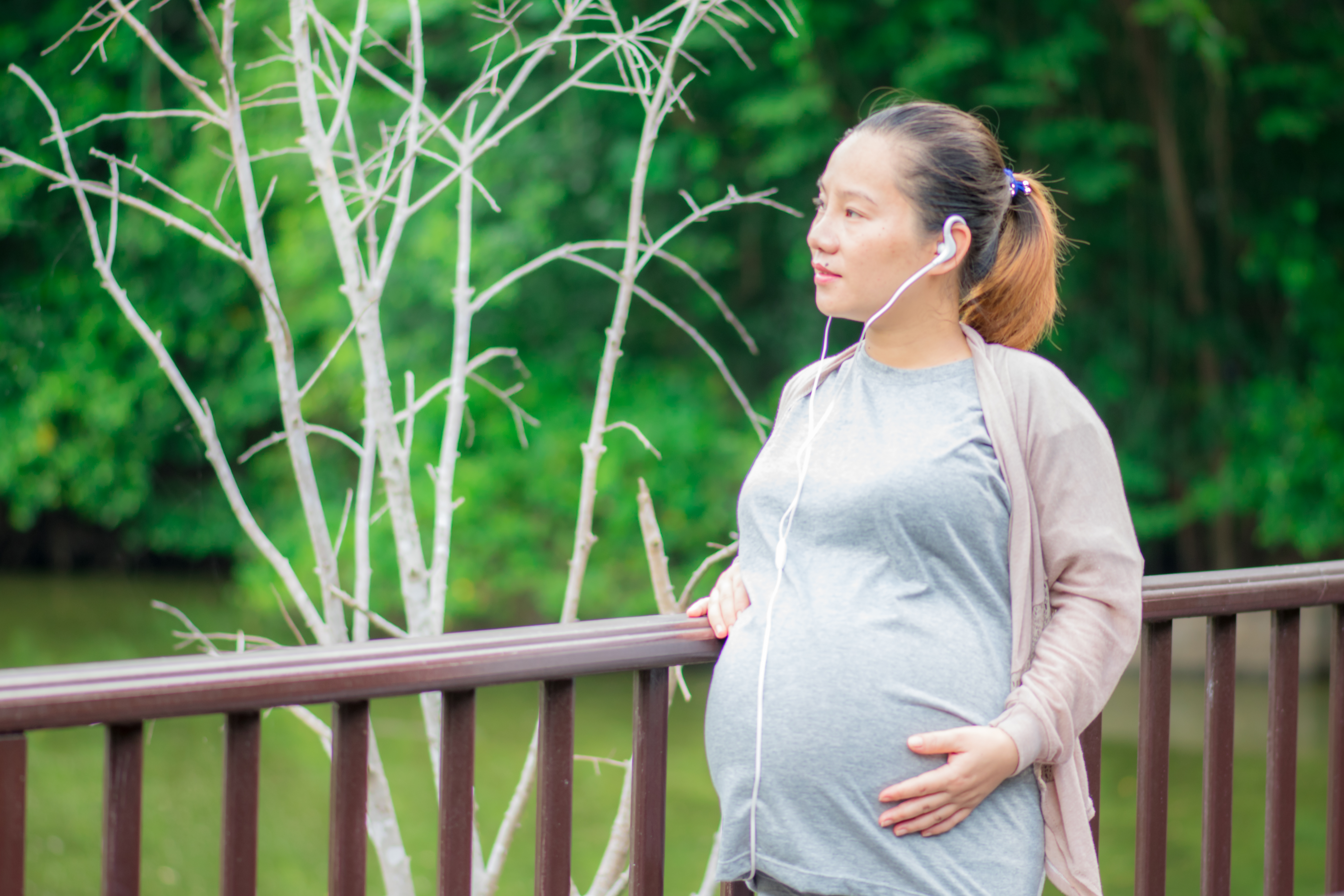 Pregnant woman with earphones leans against a railing beside a river