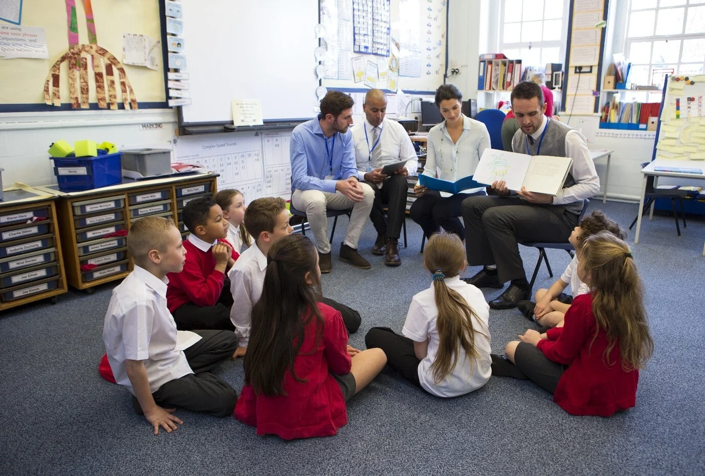 Students sitting on the floor around the teachers who are holding an open book