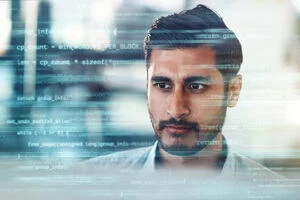 A man in a shirt sits on a computer in an office setting with code surrounding him.