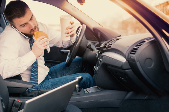 Man in car eating food, talking on phone and using laptop computer.