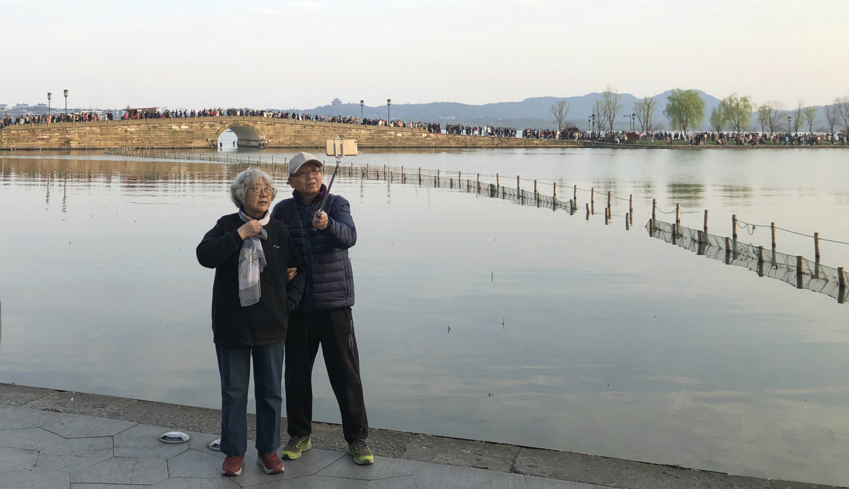 Older couple using a selfie stick to take a selfie in front of a lake in Shanghai. They are dressed for winter and both looking at the smartphone.