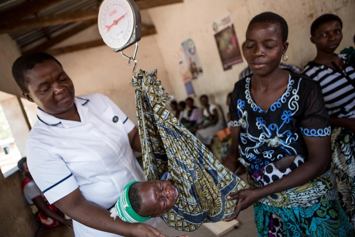 A female health care worker weighs a baby using scales as the baby's mother looks on