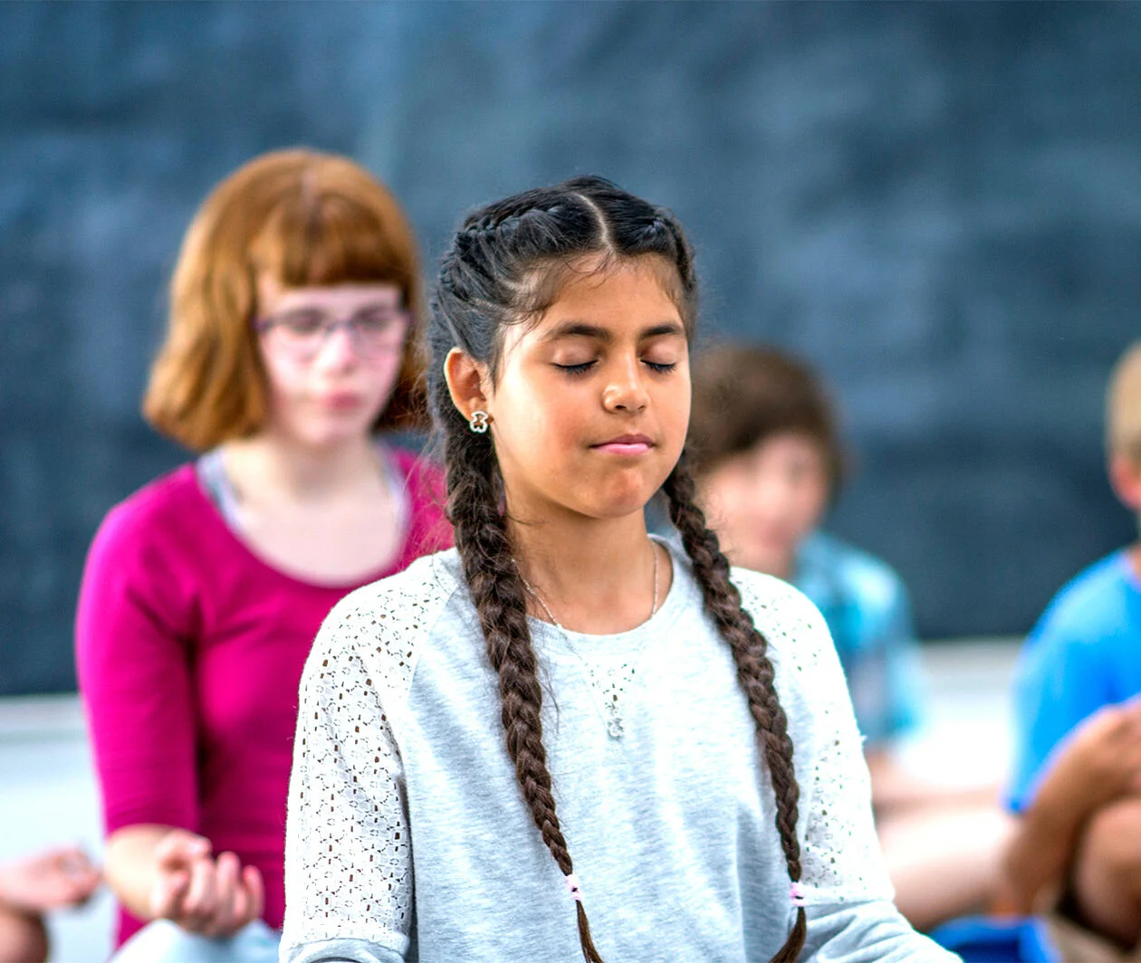 A young girl meditates in a classroom with other students.