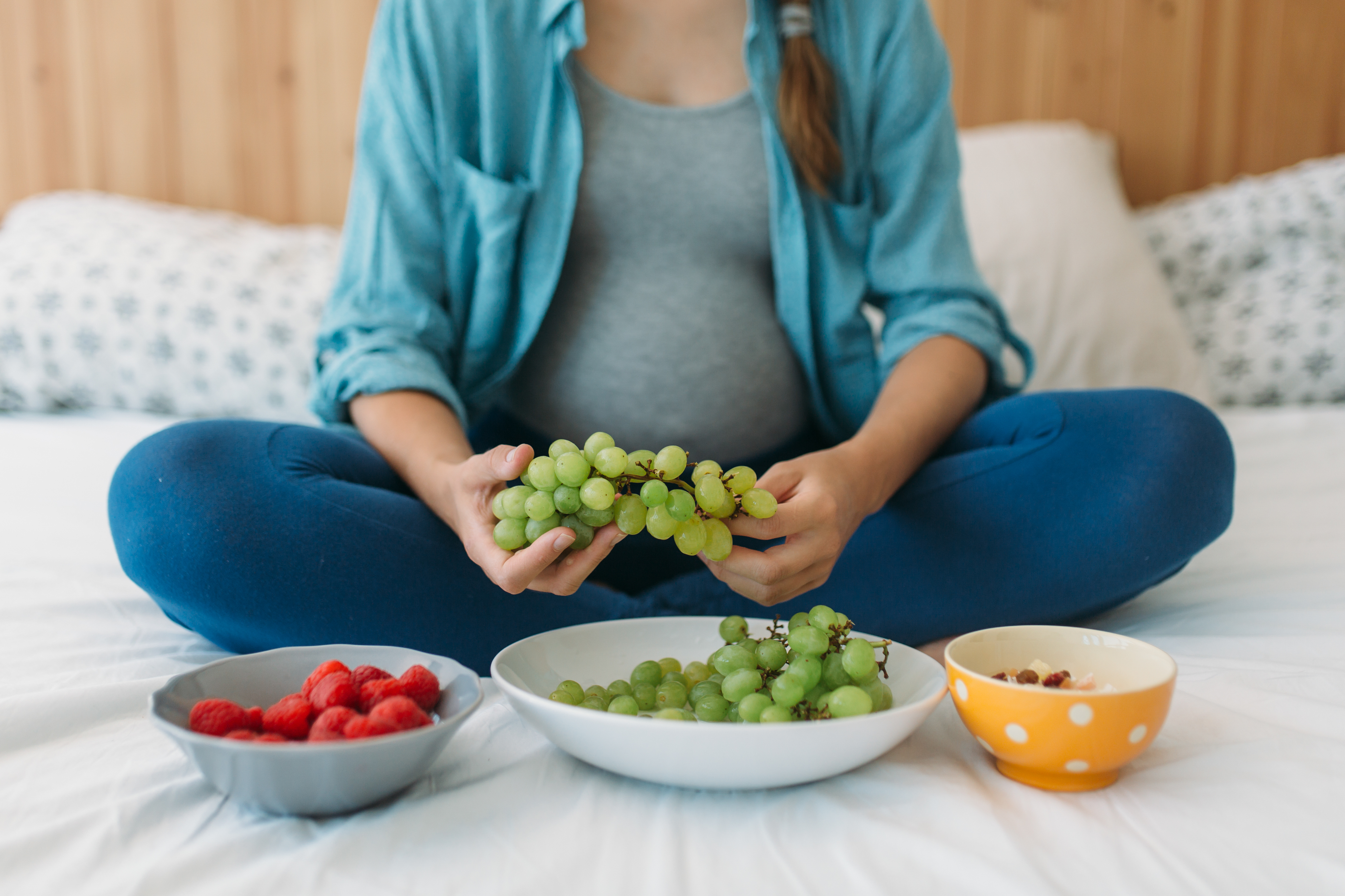 Pregnant woman holding grapes and sitting on bed next to three bowls of fruit.