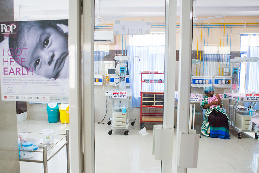 Looking through the doors to the NICU (with an ROP health awareness poster on the wall) at a mother tenderly holding her baby beside the incubator