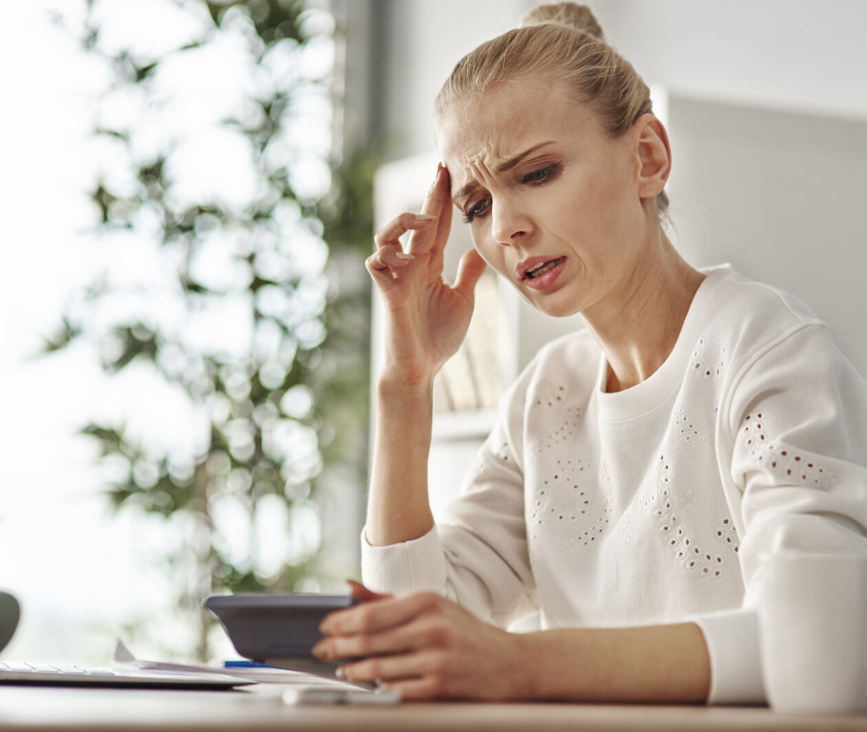 Image of a female sitting at a table looking worried and holding a calculator in one hand and the other hand is holding her head.