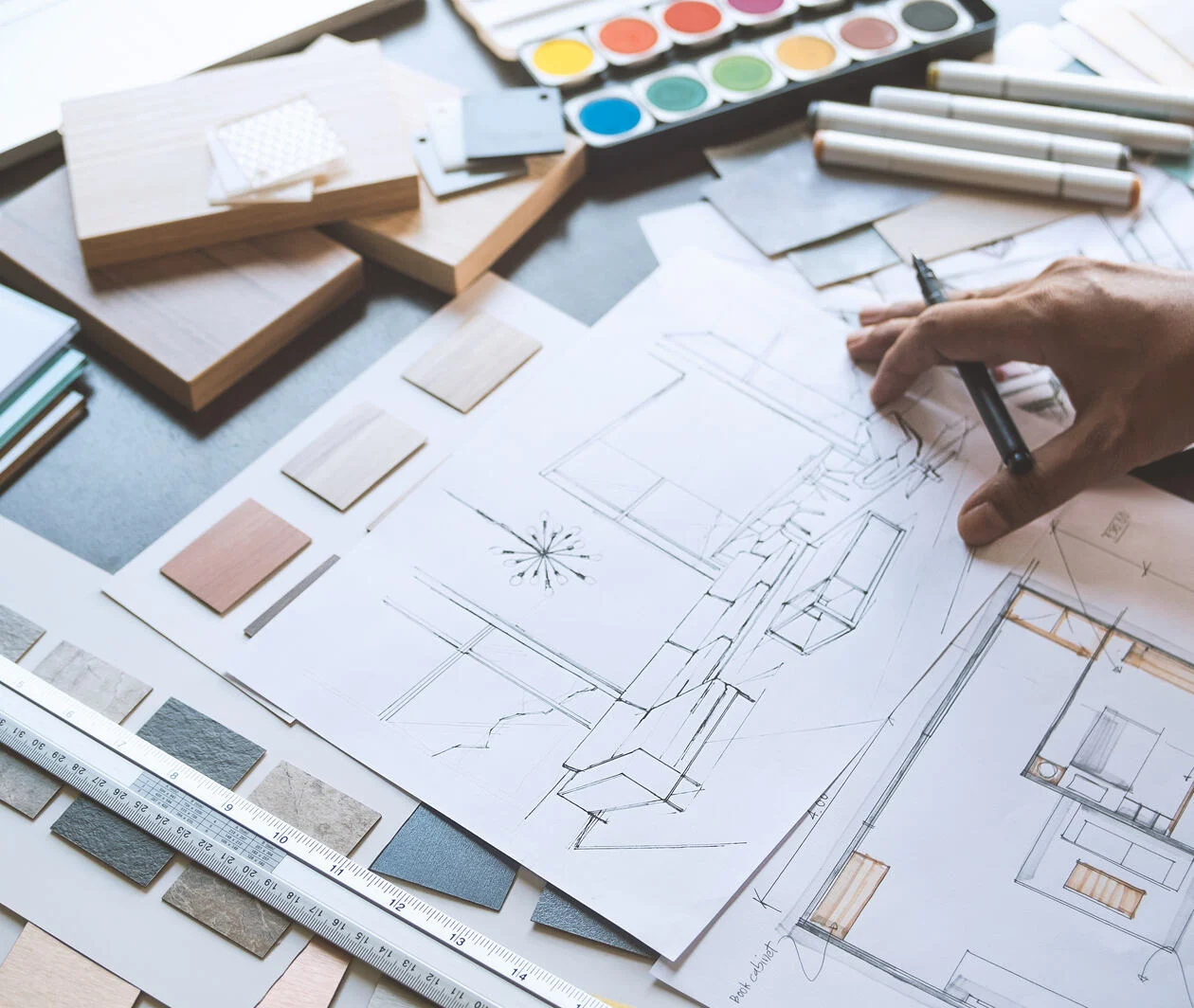 An Interior Designer's desk on top of which are drawings of a living space, samples for colours and furniture finishes as well as different coloured paints and pens. 