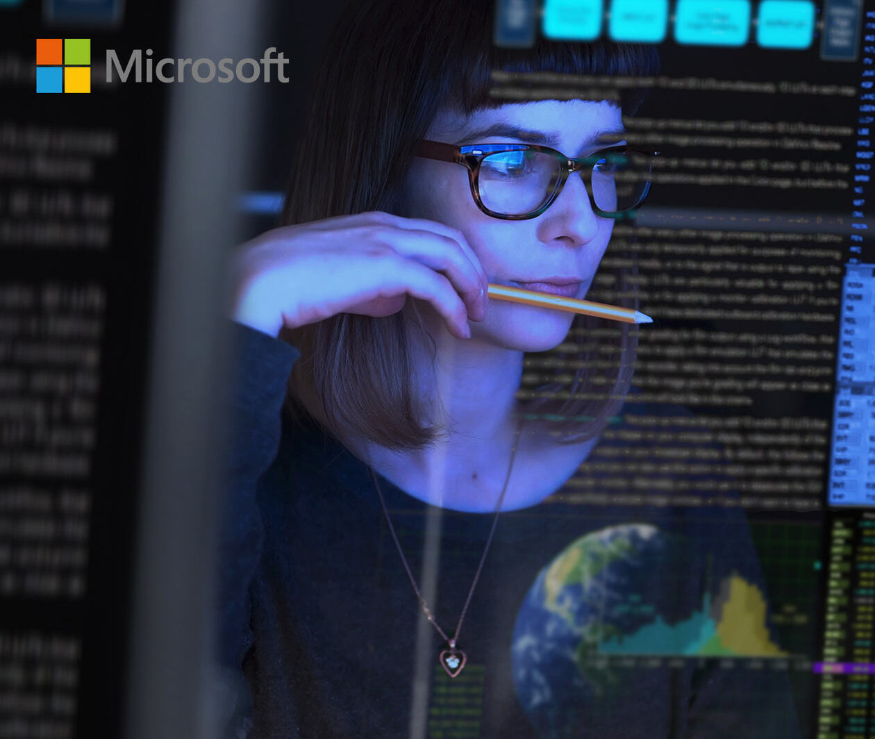 A woman with glasses sits gazing at her computer screen surrounded by code, whilst holding a pencil. 