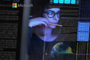 A woman with glasses sits gazing at her computer screen surrounded by code, whilst holding a pencil. 