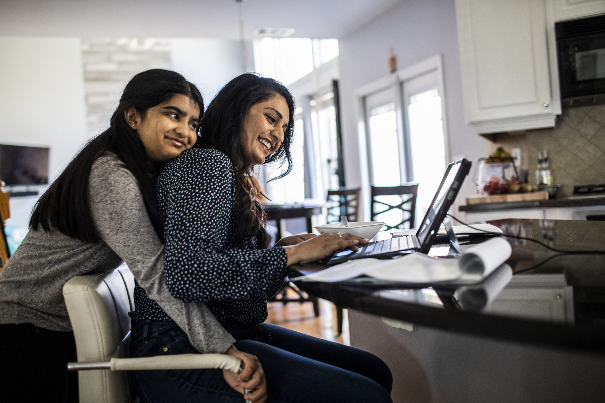 A mother at a desk working on a laptop whilst her daughter hugs her from behind.