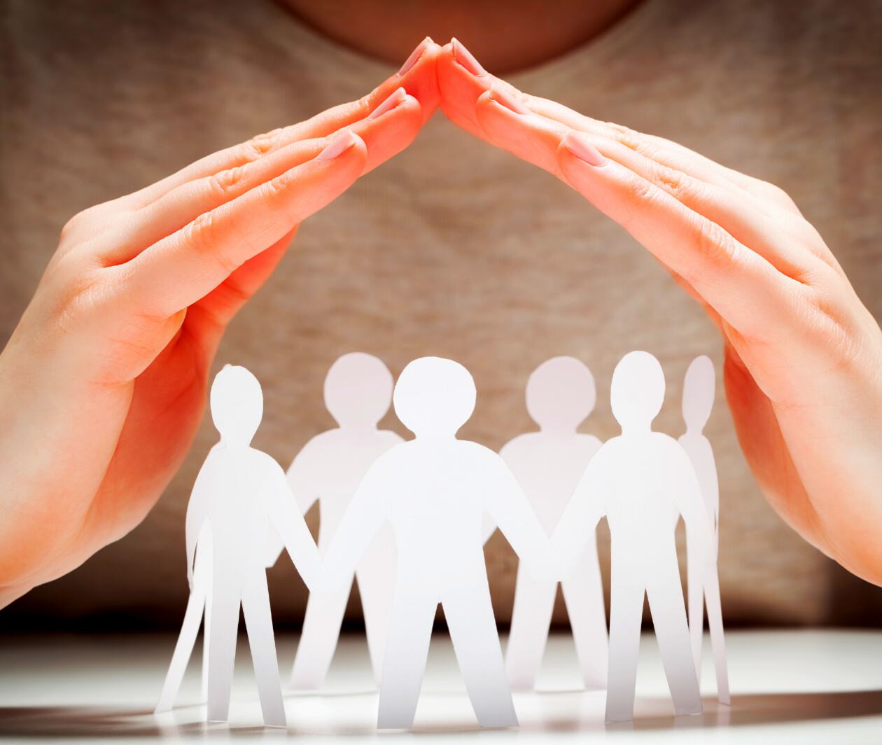 A pair of hands raised with fingers touching to form a cover of protection above a circle of cut out paper dolls. 