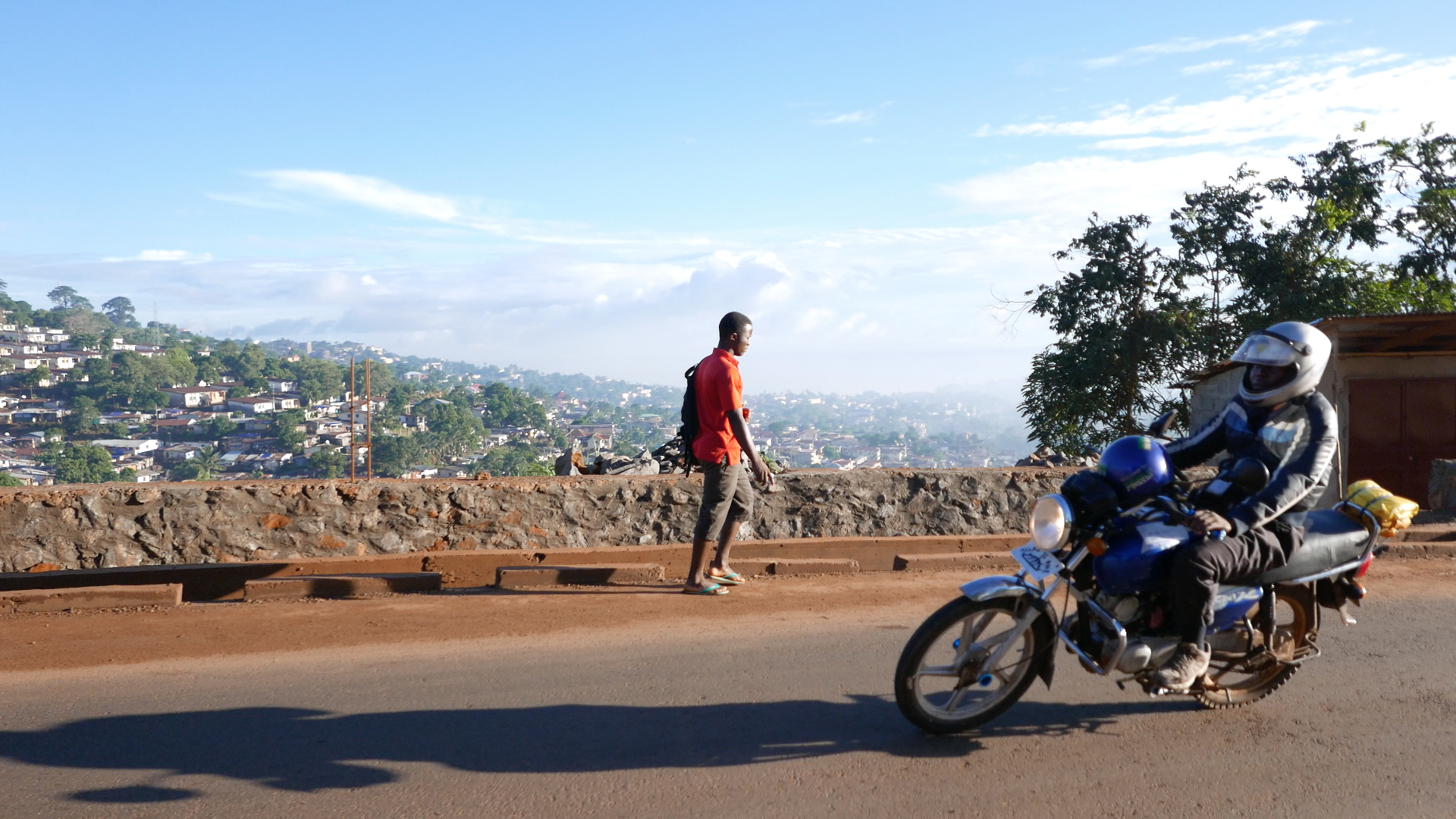 A motorbike passing a person on foot with a view over Freetown in the background