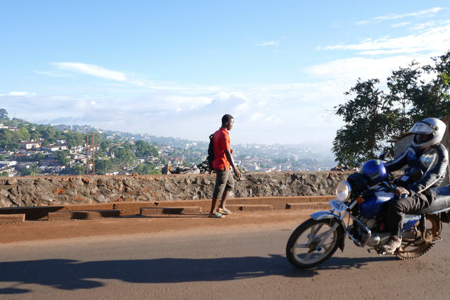 A motorbike passing a person on foot with a view over Freetown in the background