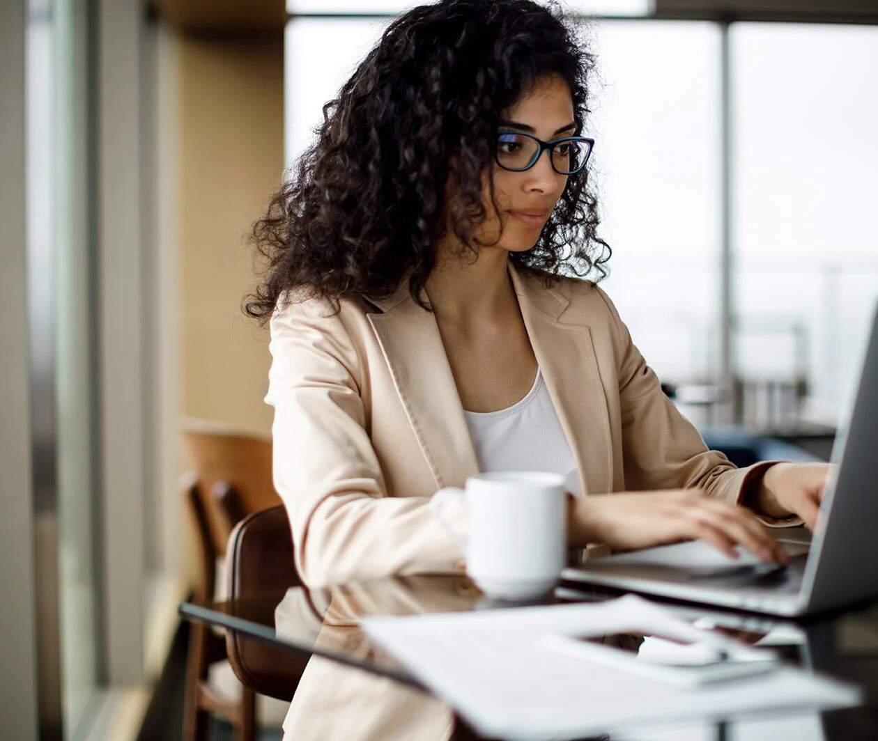 A woman in a beige blazer and glasses sits on a laptop in an office setting.