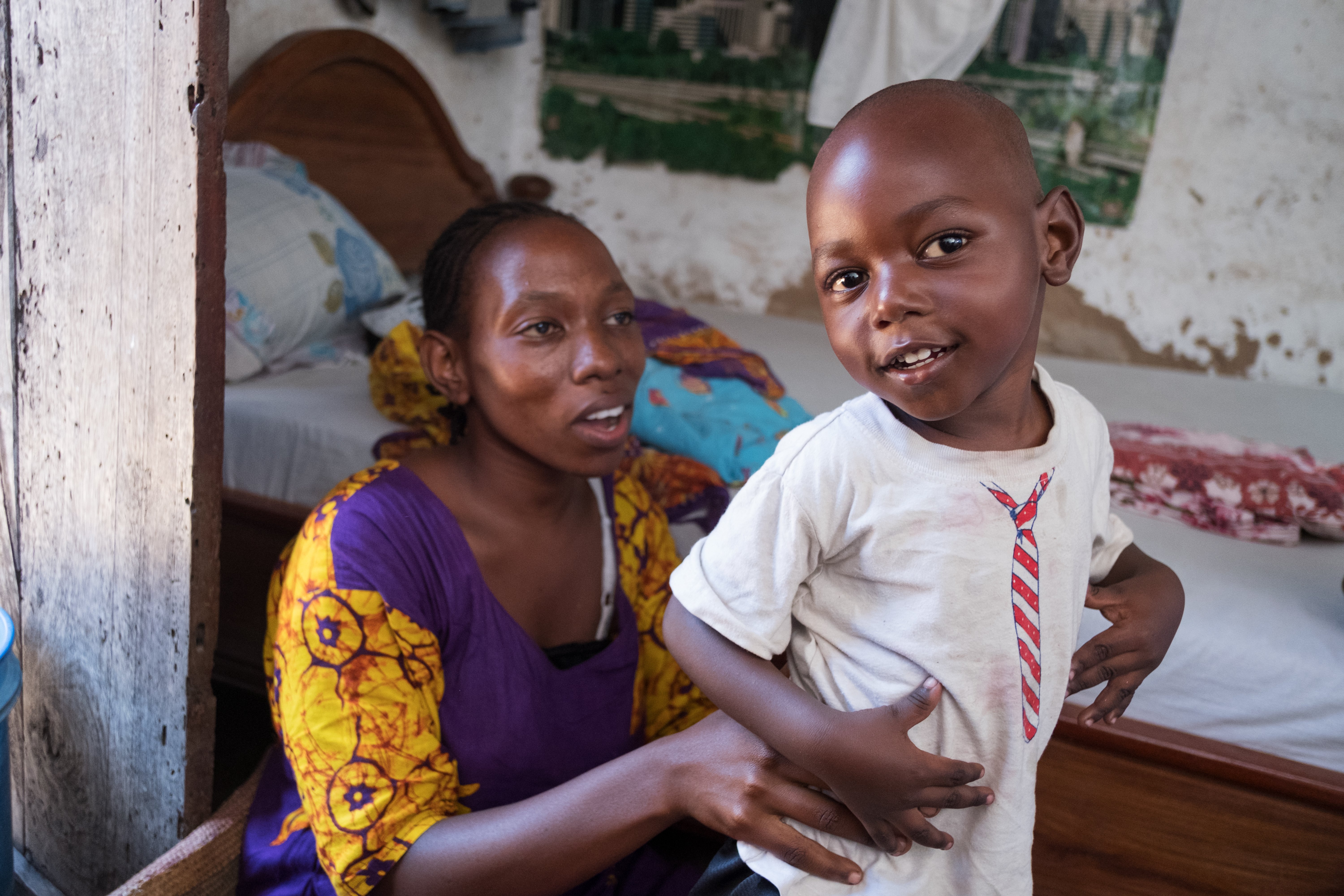 A mother helps support her son to stand whilst in his bedroom.