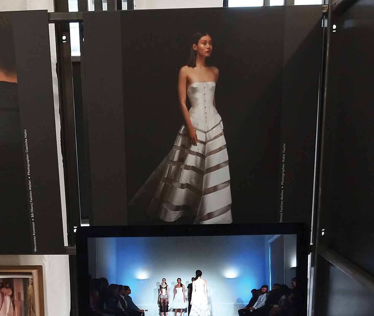 A photograph of a model posing wearing a white garment with a laptop on the table in front.