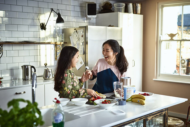 Woman holding yoghurt on spoon towards girl, laughing, threatening to smear food on daughter's face, messing about, having fun, playing, bonding