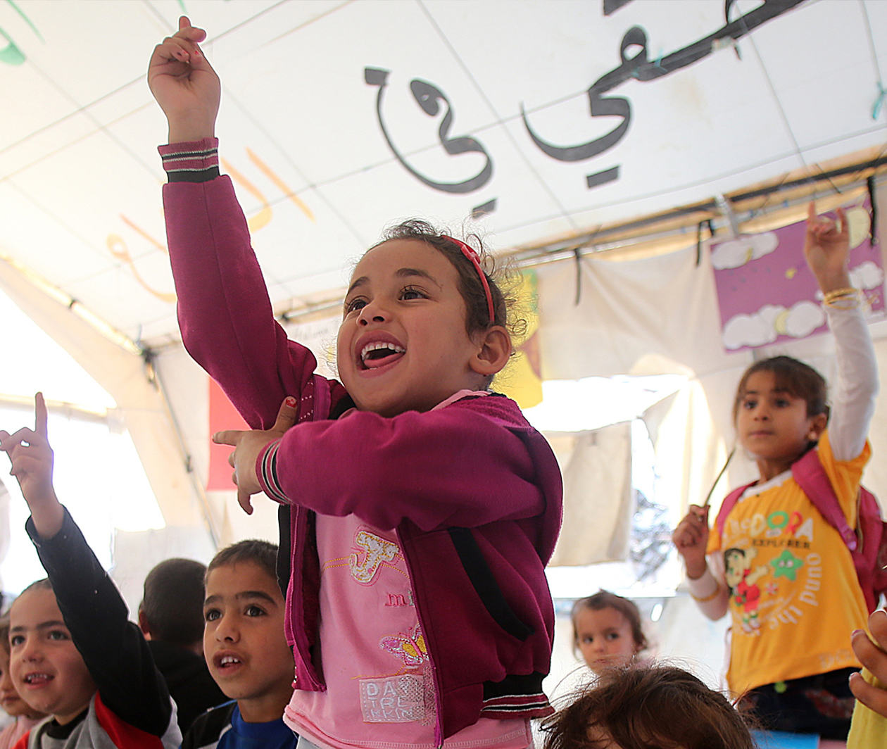 children in a makeshift classroom putting up their hands to answer a question