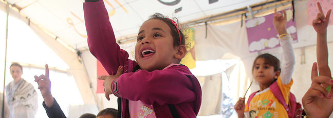 children in a makeshift classroom putting up their hands to answer a question