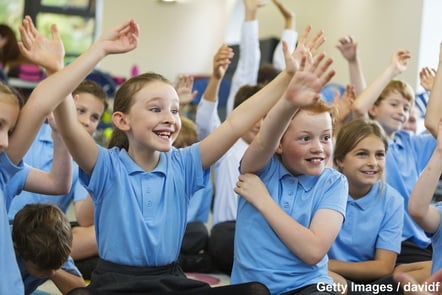 A group of excited-looking pupils in a classroom, most of whom are facing the front and have either one arm or both arms raised.