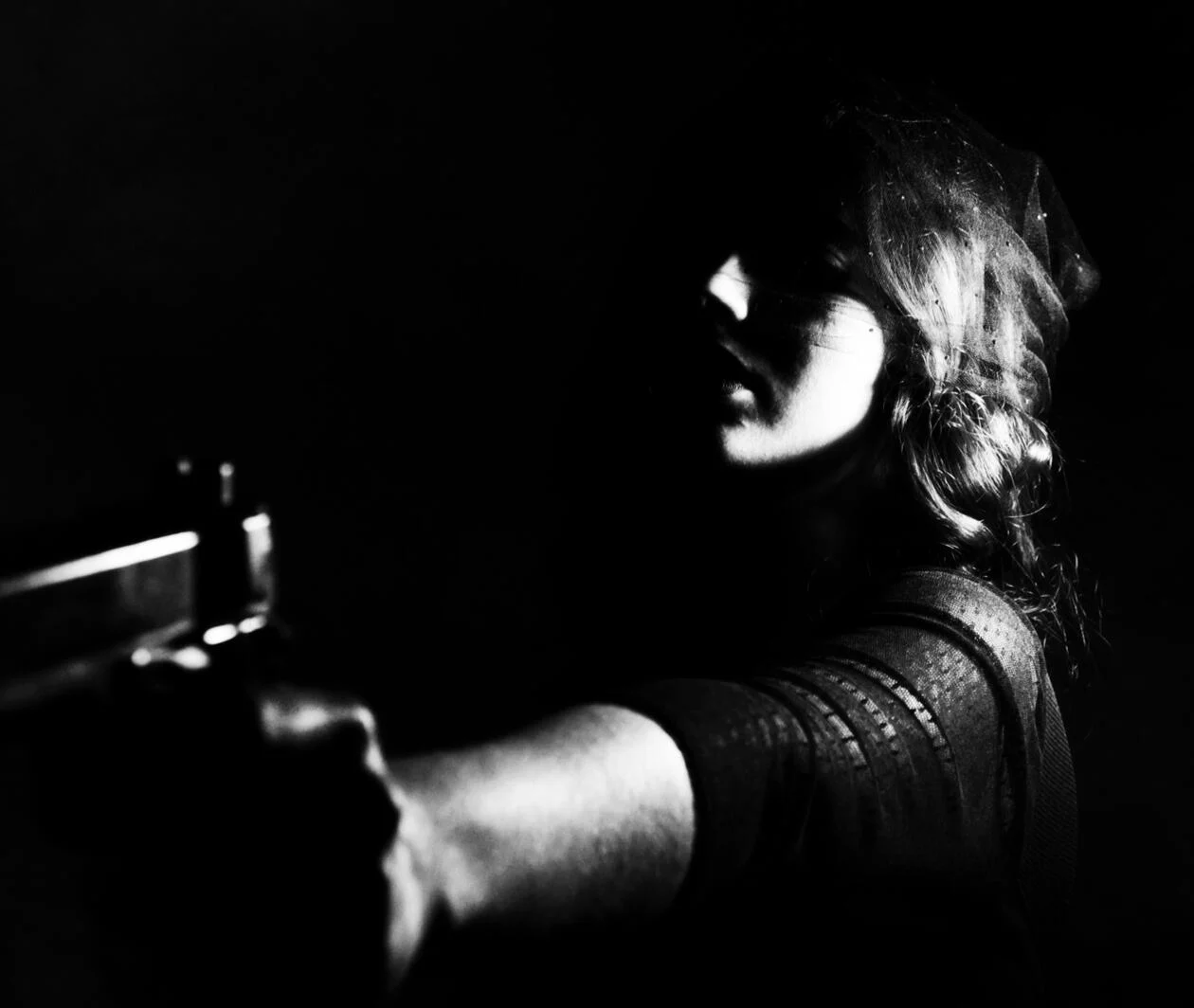 A young woman peering from the shadows holding a handgun