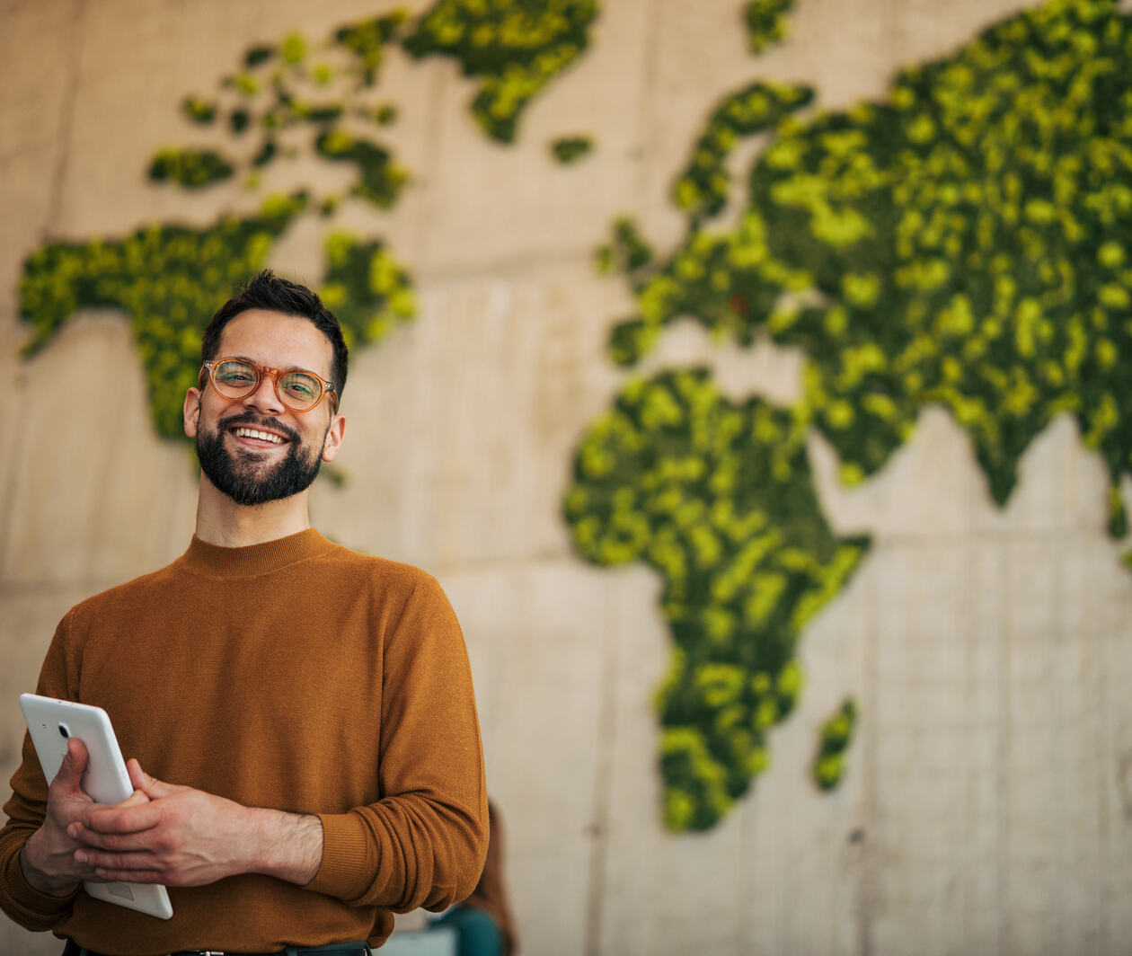 Happy man holding a digital tablet smiling at the camera, promoting global sustainability, technology, and environmental responsibility with a moss world map in the background