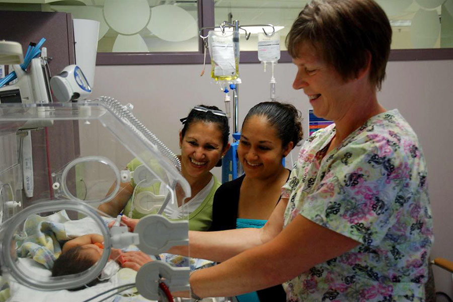 Mother and grandmother smiling at a preterm baby inside an incubator as a smiling member of the neonatal team checks their baby