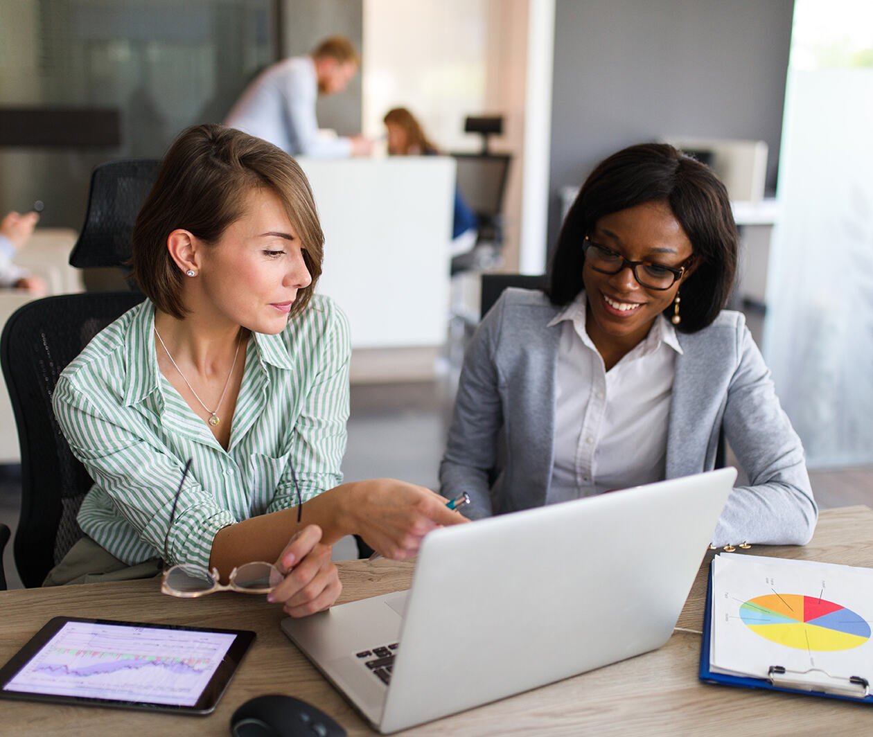 Two businesswomen are in the office using laptop, digital tablet and chart papers while working together.