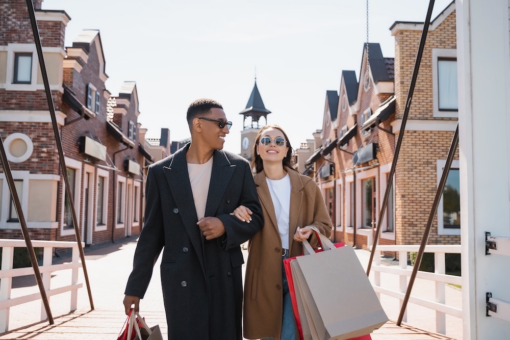 Front of view of man and woman linking arms holding shopping bags smiling with houses in background