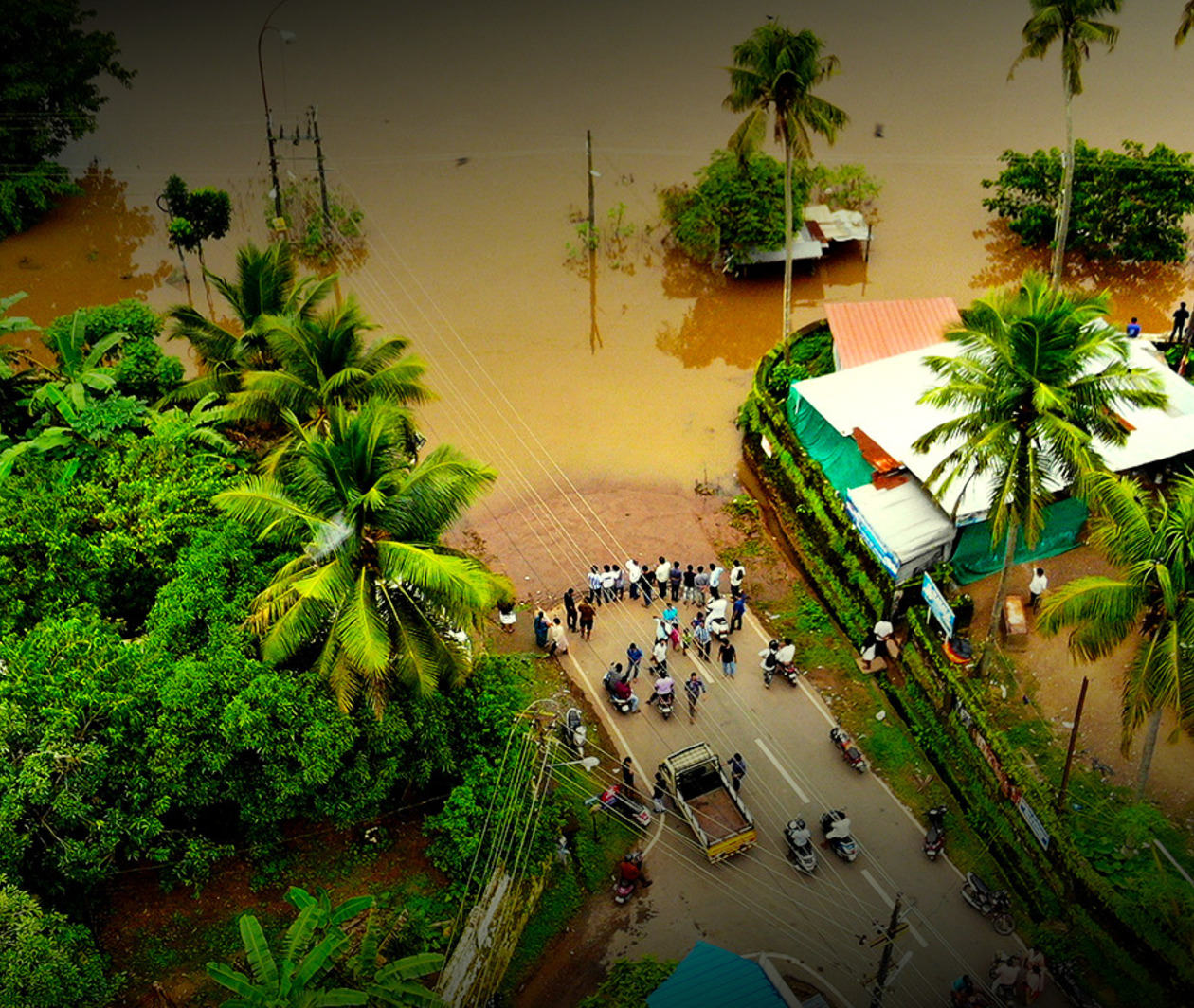 An aerial photograph of a flooded village in Kerala, India - villagers stand on a road cut off by water