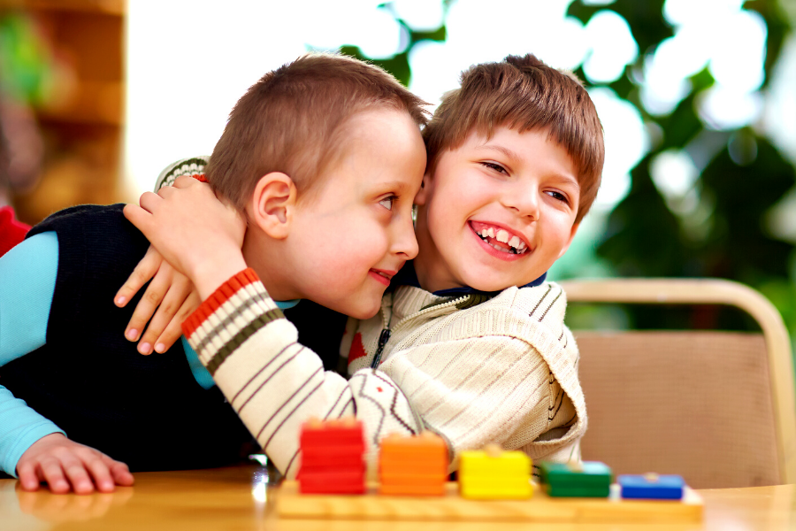 Two children with disabilities are seated at a table, hugging. One child is smiling towards the camera. The other child's face is extremely close to the first child and their open eyes are staring at the first child's cheek.