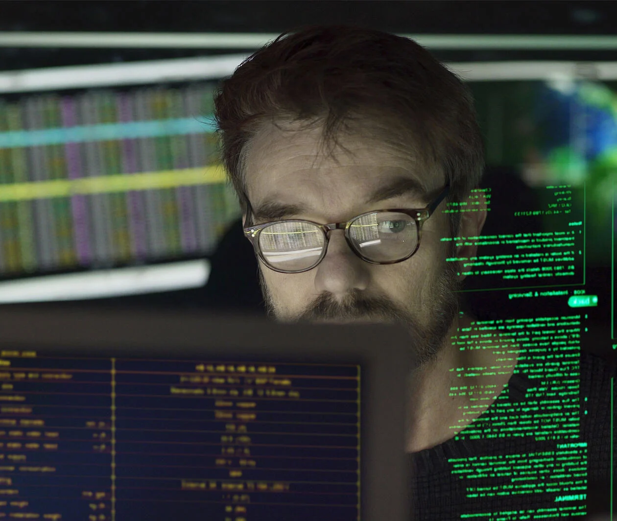 A man with glasses looking at multiple computer screens. 