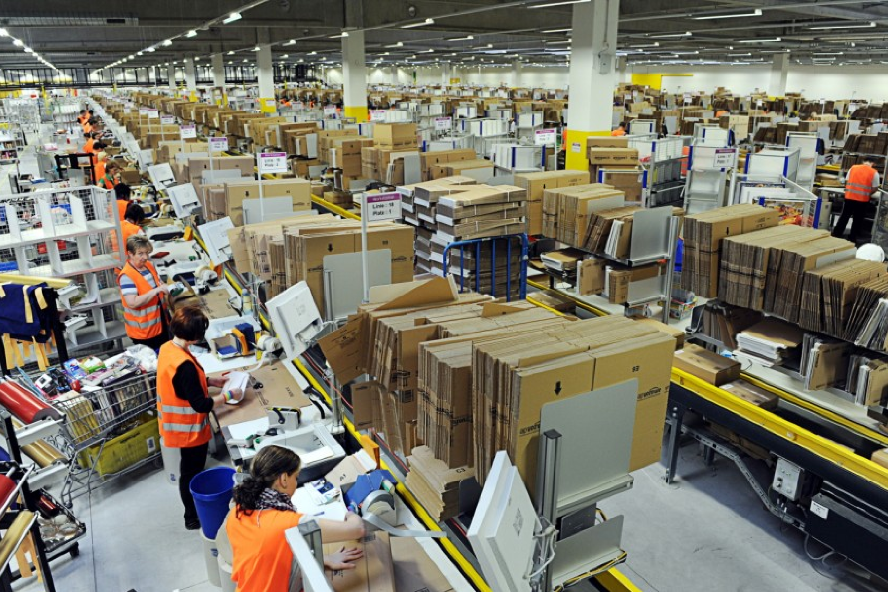 a photograph of workers on the production line in an Amazon warehouse. There's entire image is filled with rows of boxes and packages.