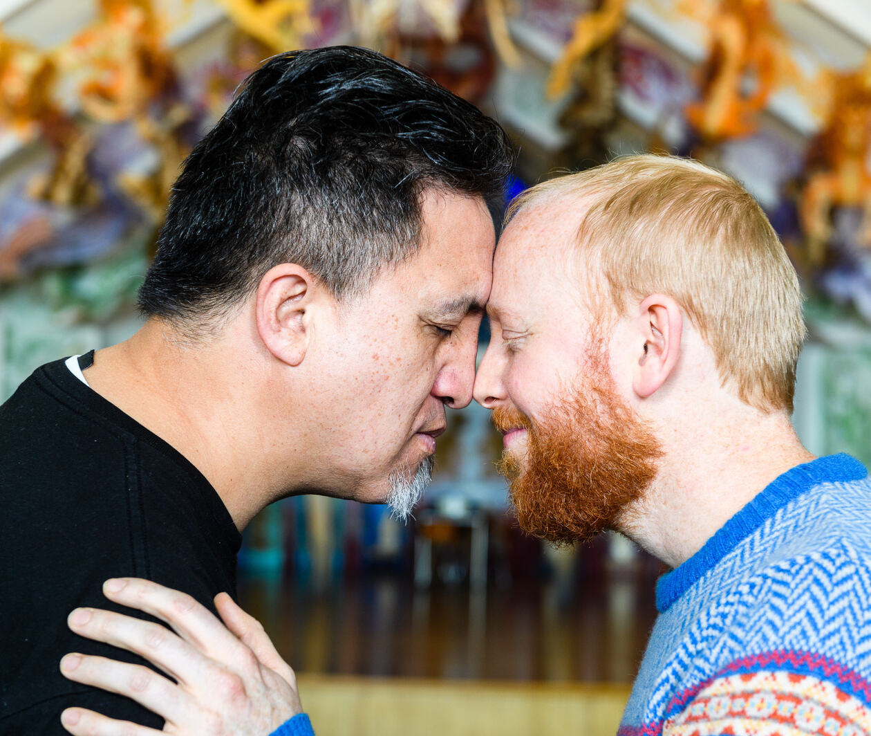 Two men stand in profile, pressing their noses together in a traditional Māori hongi. The man on the left is Māori with dark hair and brown skin. The man on the right is European with light skin and ginger hair and beard. They stand in front of a marae.