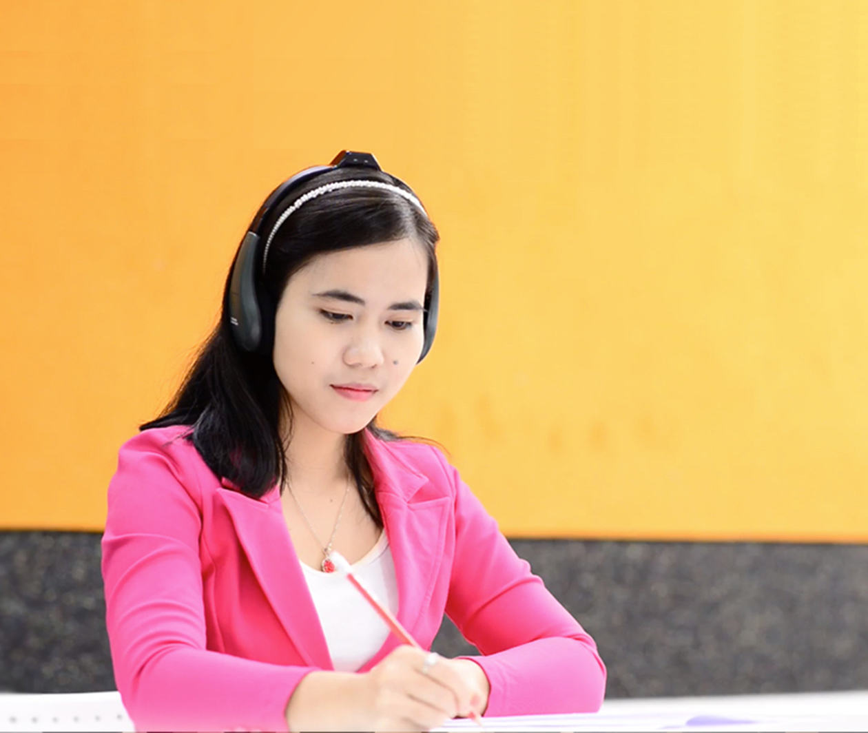 Woman sat at a table with headphones on with pencil in hand writing on paper