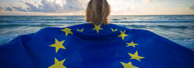 Woman walking on beach towards the sea and sun as it emerges from the clouds with the European Union flag draped around her shoulders. 