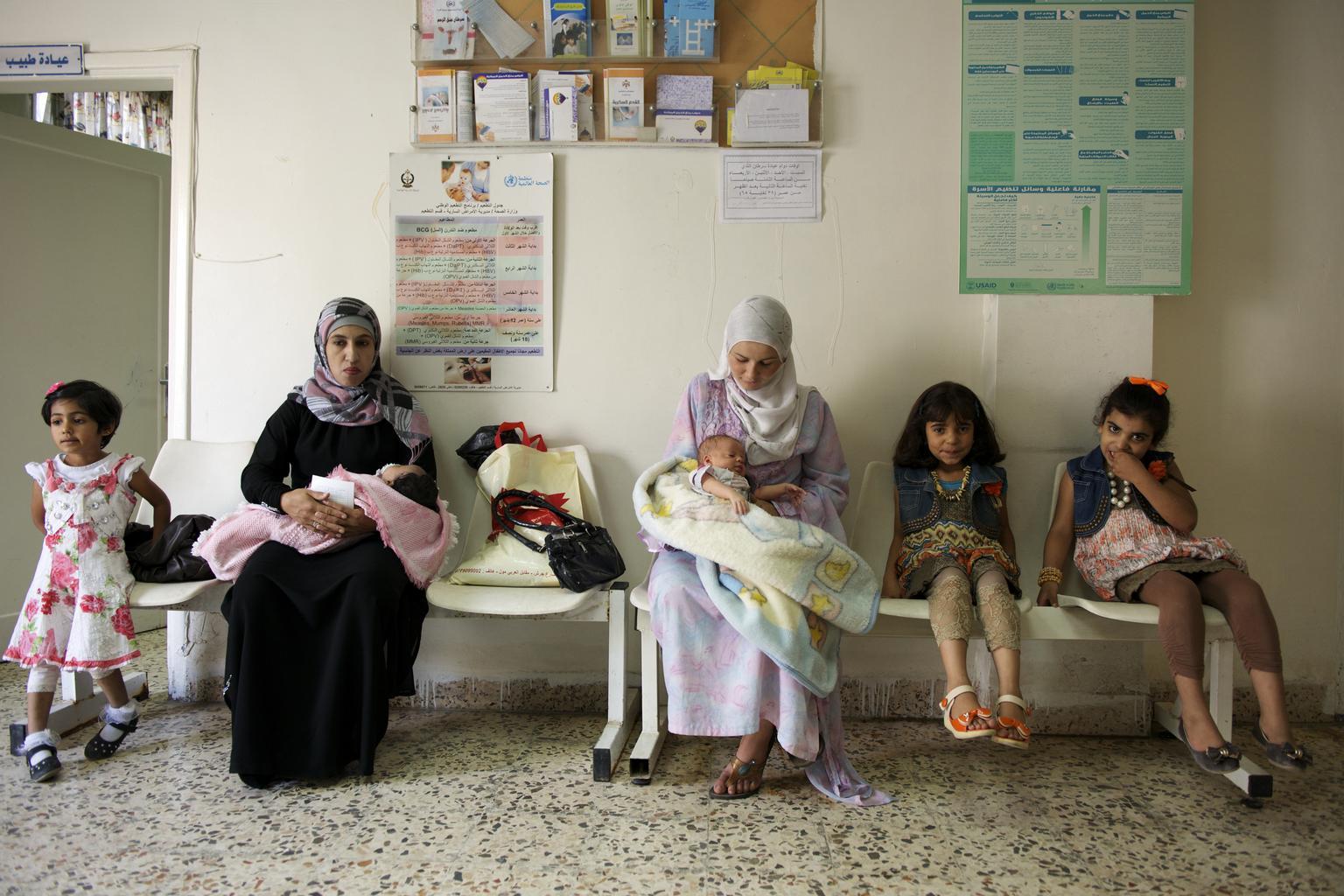 Two mothers holding their babies are shown sitting in a waiting room. Other small children sit on chairs nearby.