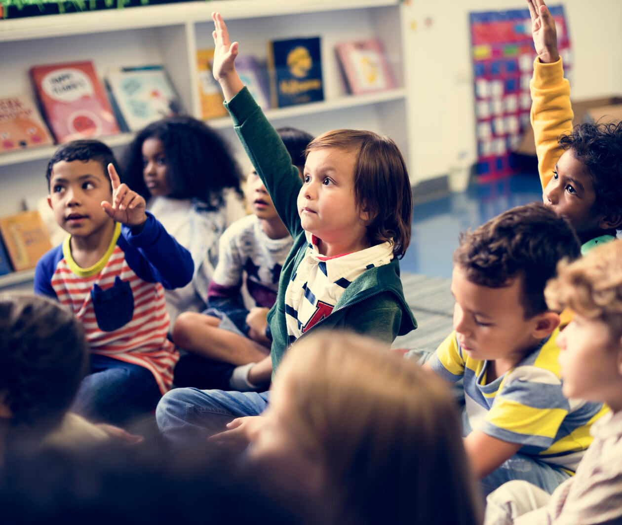 Photo of a group of children sitting on the floor in a classroom, looking n one direction, several with their hands up.